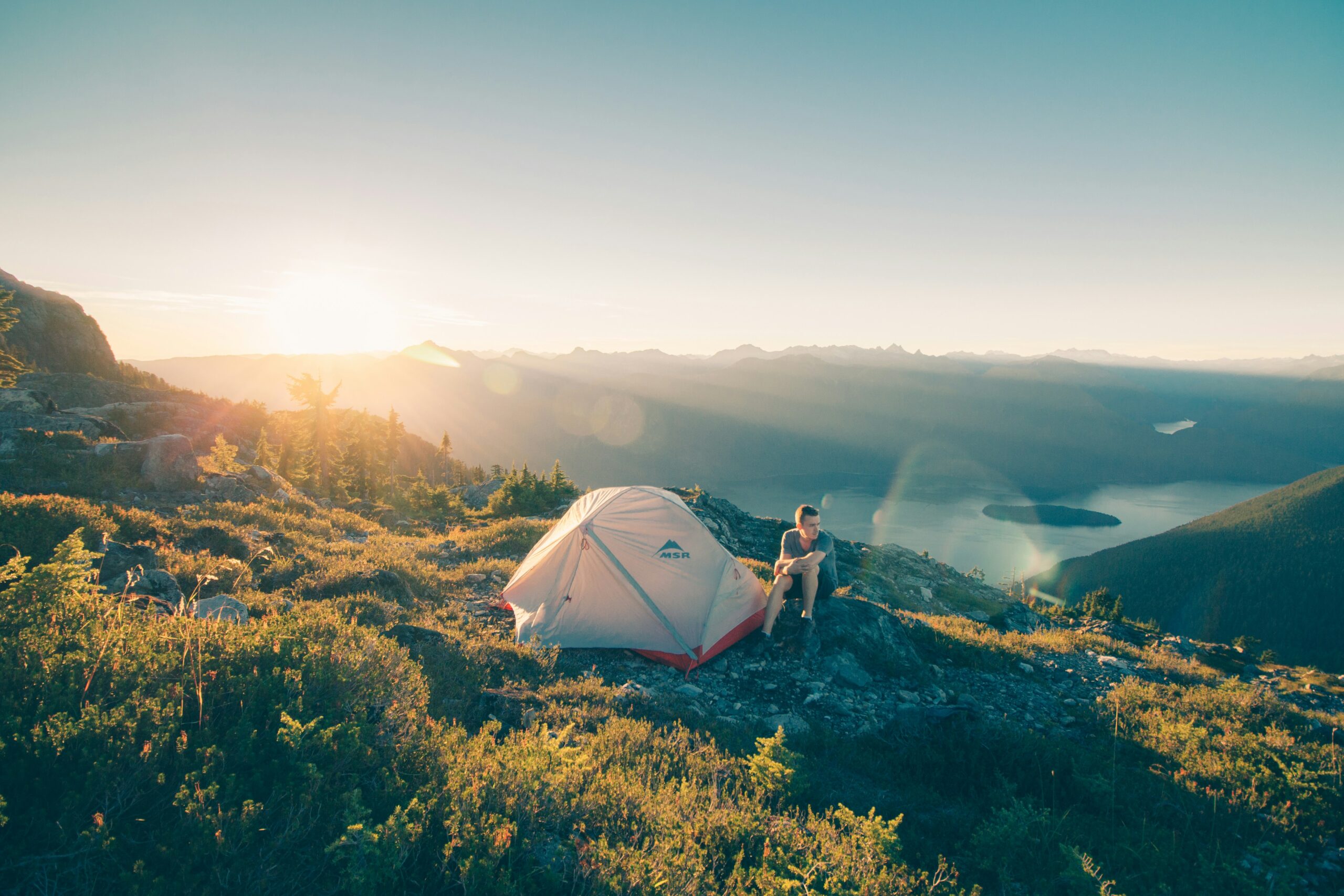Man camping with a tent on a mountain ridge overlooking a lake at sunrise.